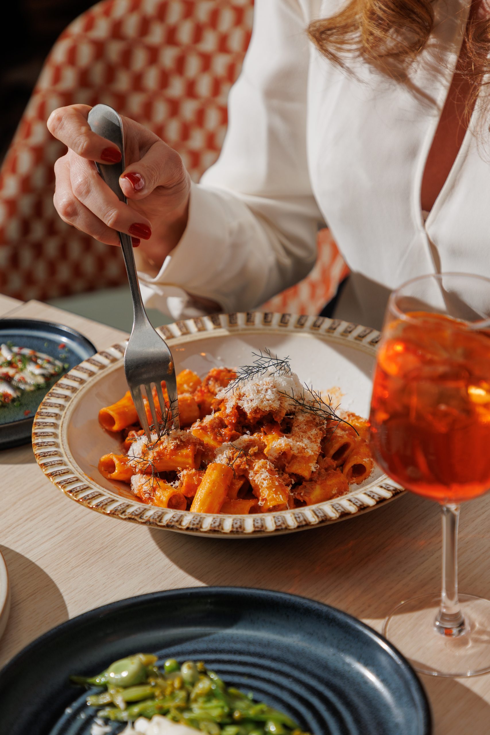 Woman in white blouse eating pasta at a restaurant table with an Aperol Spritz.